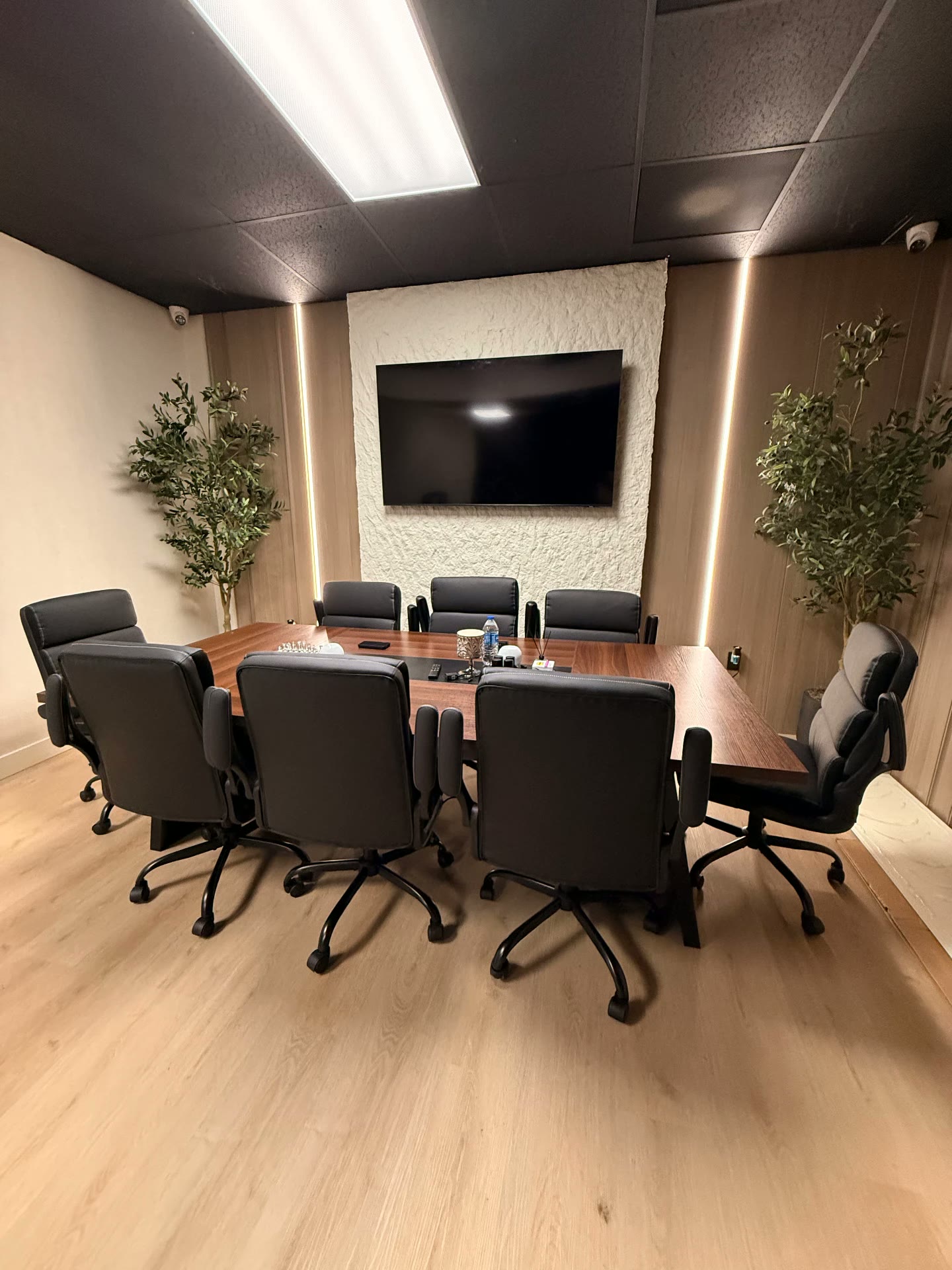 Sala de Juntas — Comercial · Oficina Sala de juntas corporativa con mesa de madera nogal, ocho sillas ejecutivas negras, paneles de madera con iluminación LED vertical, muro texturizado con pantalla y olivos decorativos