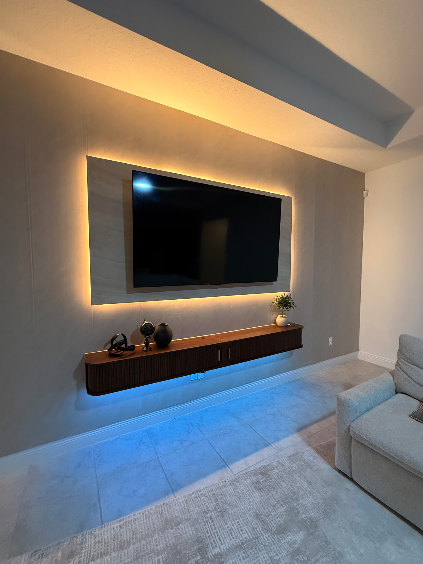 Muro TV Retroiluminado — Residencial · Sala de TV Muro de TV en microcemento con marco de porcelanato retroiluminado en luz cálida, mueble flotante acanalado de nogal con luz LED azul y sillón gris claro