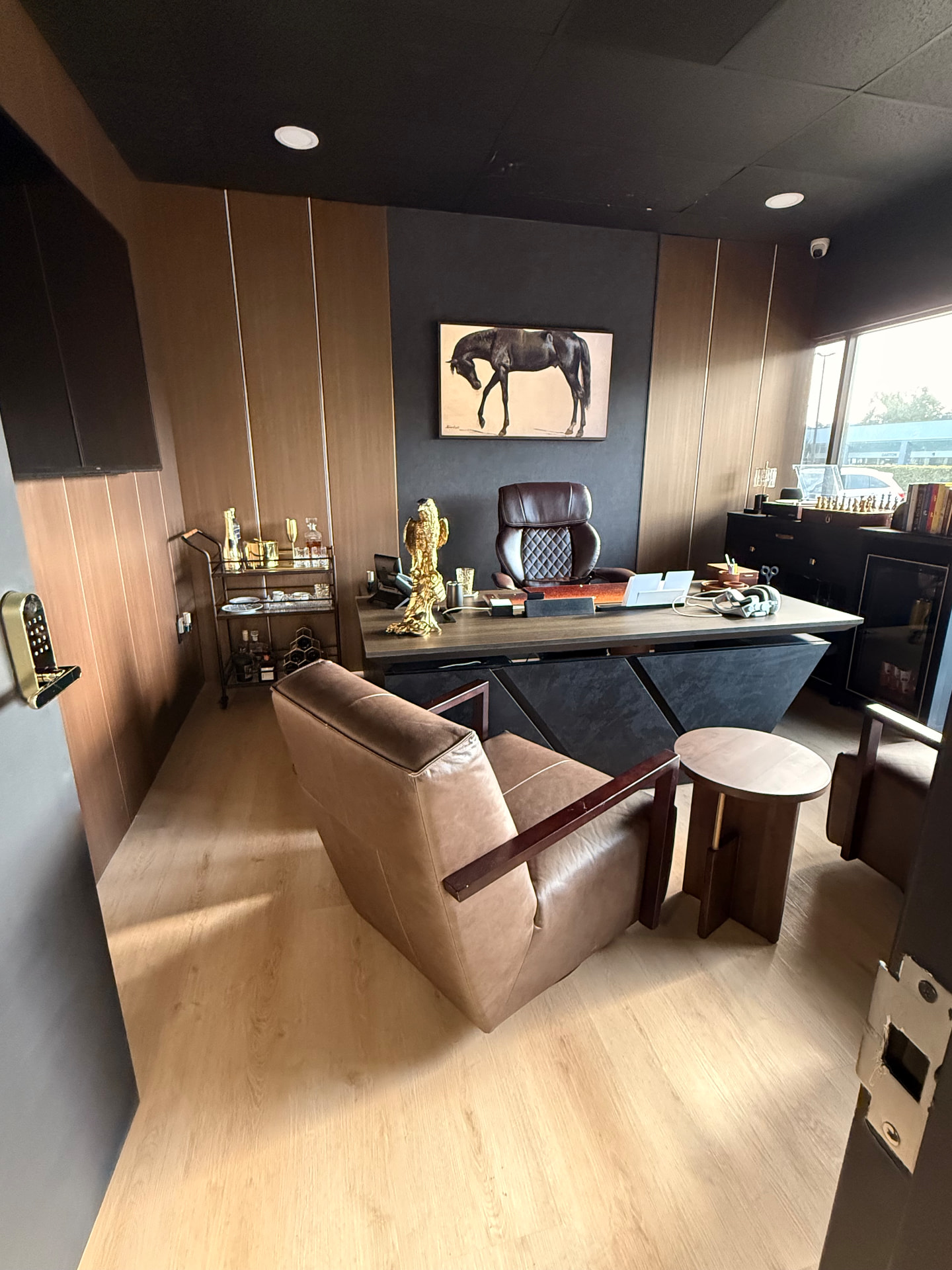 Oficina Ejecutiva — Comercial · Oficina privada Oficina ejecutiva con paneles de madera roble, techo negro, escritorio flotante gris, sillón de cuero, carrito de bar dorado y cuadro de caballo sobre muro texturizado oscuro