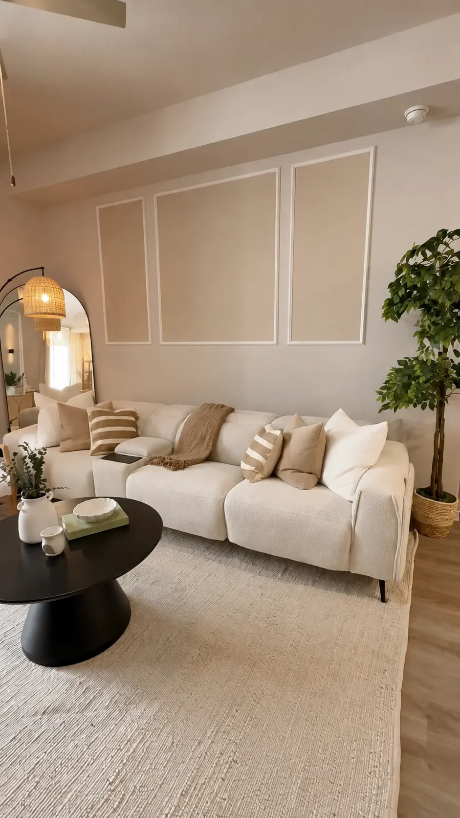 Sala Serena — Residencial · Sala Sala residencial en tonos crema y beige con sofá modular blanco, cojines de lino y rayas, molduras decorativas en la pared, alfombra de yute, mesa de centro negra redonda y planta de interior